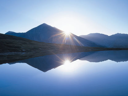 Bright sunrise over a mountain lake, with sun rays reflecting perfectly on the still water, by John Fielder.