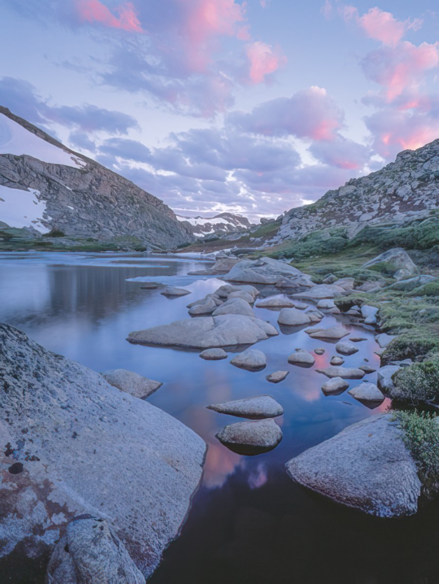 Tranquil mountain lake at dusk, surrounded by rocks and snow-dusted peaks under a pink and blue clouded sky, captured by John Fielder.