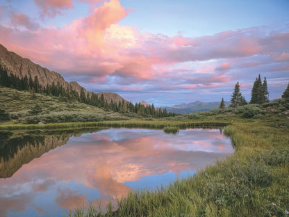 Expansive mountain landscape with a calm lake reflecting pink and orange clouds, framed by green trees and distant peaks, John Fielder.