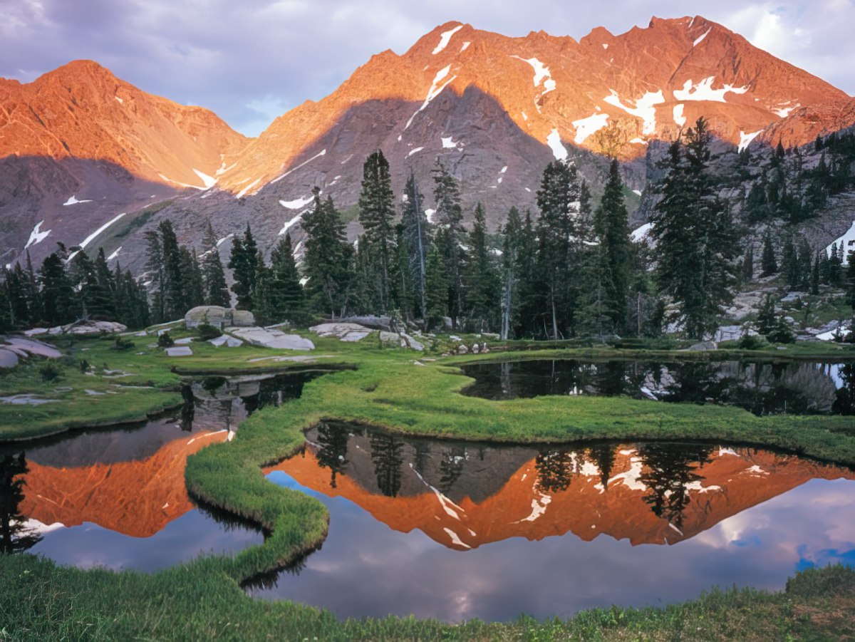 Majestic mountains with snow patches reflecting in winding streams and small ponds surrounded by vibrant green grass, by John Fielder.