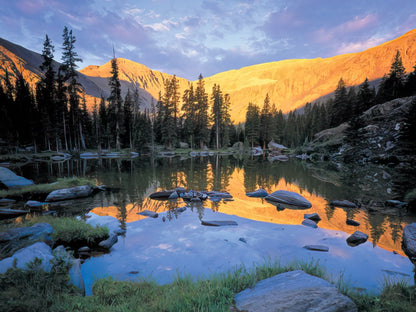 Rolling green hills with small blue lakes reflecting the sky, framed by majestic blue mountains and sparse trees, John Fielder.