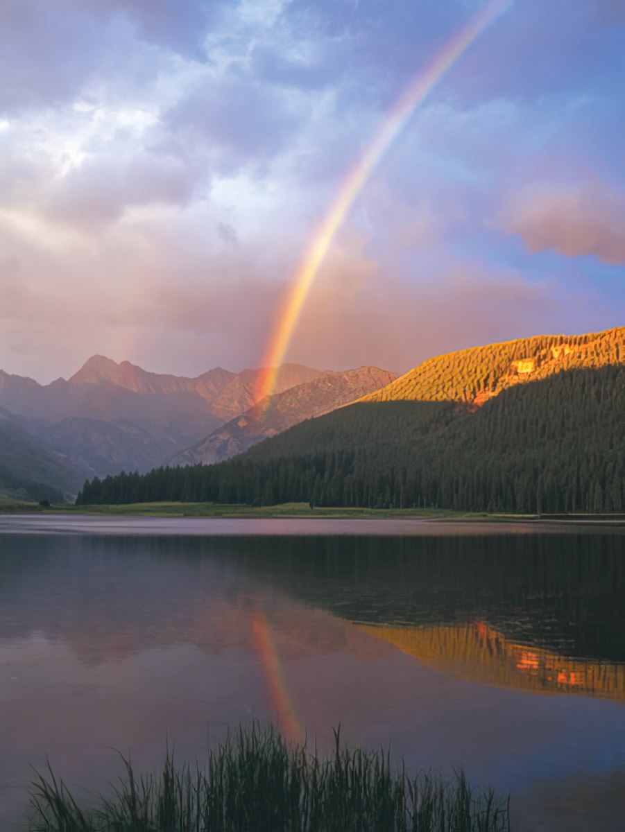 Rocky mountain lake with crystal clear water reflecting a pink cloudy sky and surrounding pine trees and snowy peaks, John Fielder.