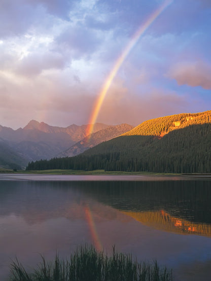 Rocky mountain lake with crystal clear water reflecting a pink cloudy sky and surrounding pine trees and snowy peaks, John Fielder.