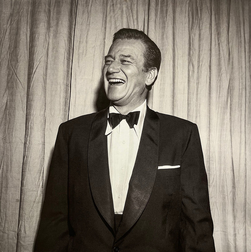 Vintage photo of John Wayne in a tuxedo, laughing heartily, featured in the John Wayne calendar.
