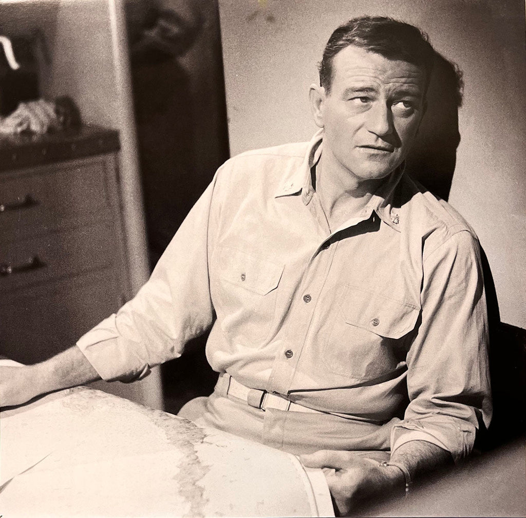Serious portrait of John Wayne in a military-style shirt, sitting at a desk with a map, vintage calendar photo.