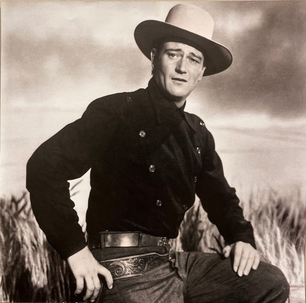 John Wayne in a black cowboy shirt, hat, and gun belt, posing in a field, a classic shot for the 2027 calendar.