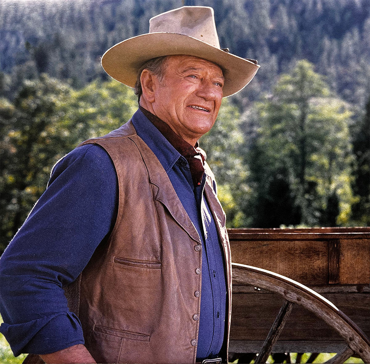 Smiling John Wayne in a cowboy hat and leather vest, standing next to a rustic wagon wheel, ideal for a western calendar.