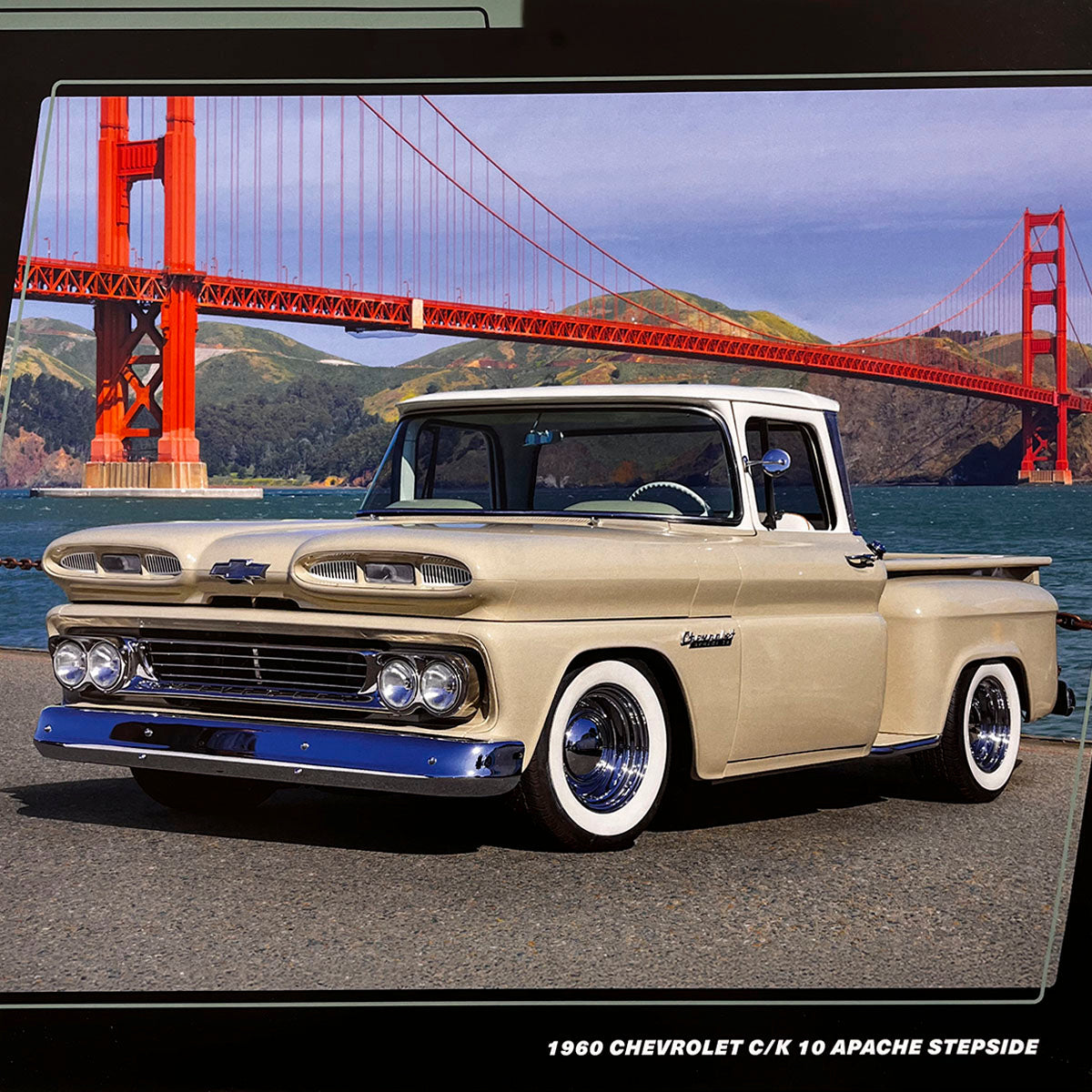 A beige 1960 Chevrolet C/K 10 Apache Stepside pickup truck with white wall tires, parked by the water with the iconic Golden Gate Bridge visible in the background under a blue sky.