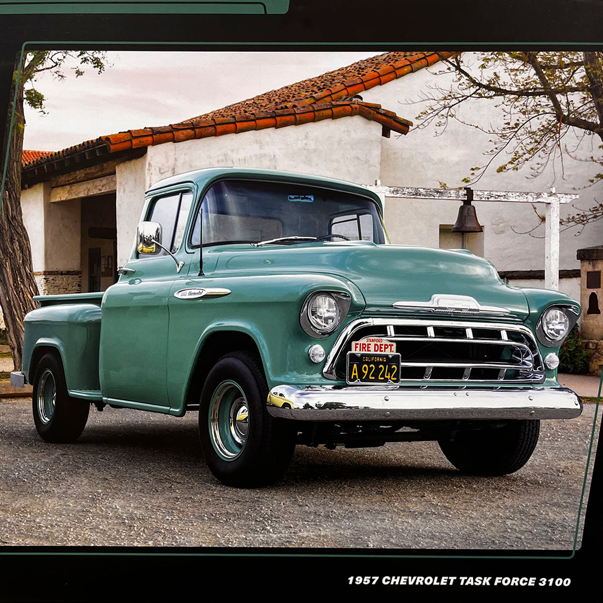 A teal green 1957 Chevrolet Task Force 3100 pickup truck with a red 'FIRE DEPT' license plate, parked beside an old adobe building, highlighting its classic utility vehicle appeal.