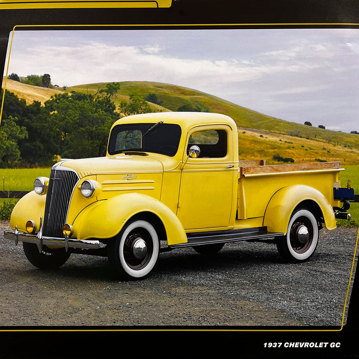 A vibrant yellow 1937 Chevrolet GC pickup truck with wooden bed rails and white wall tires, parked on a gravel path against a backdrop of rolling green hills and a clear sky.