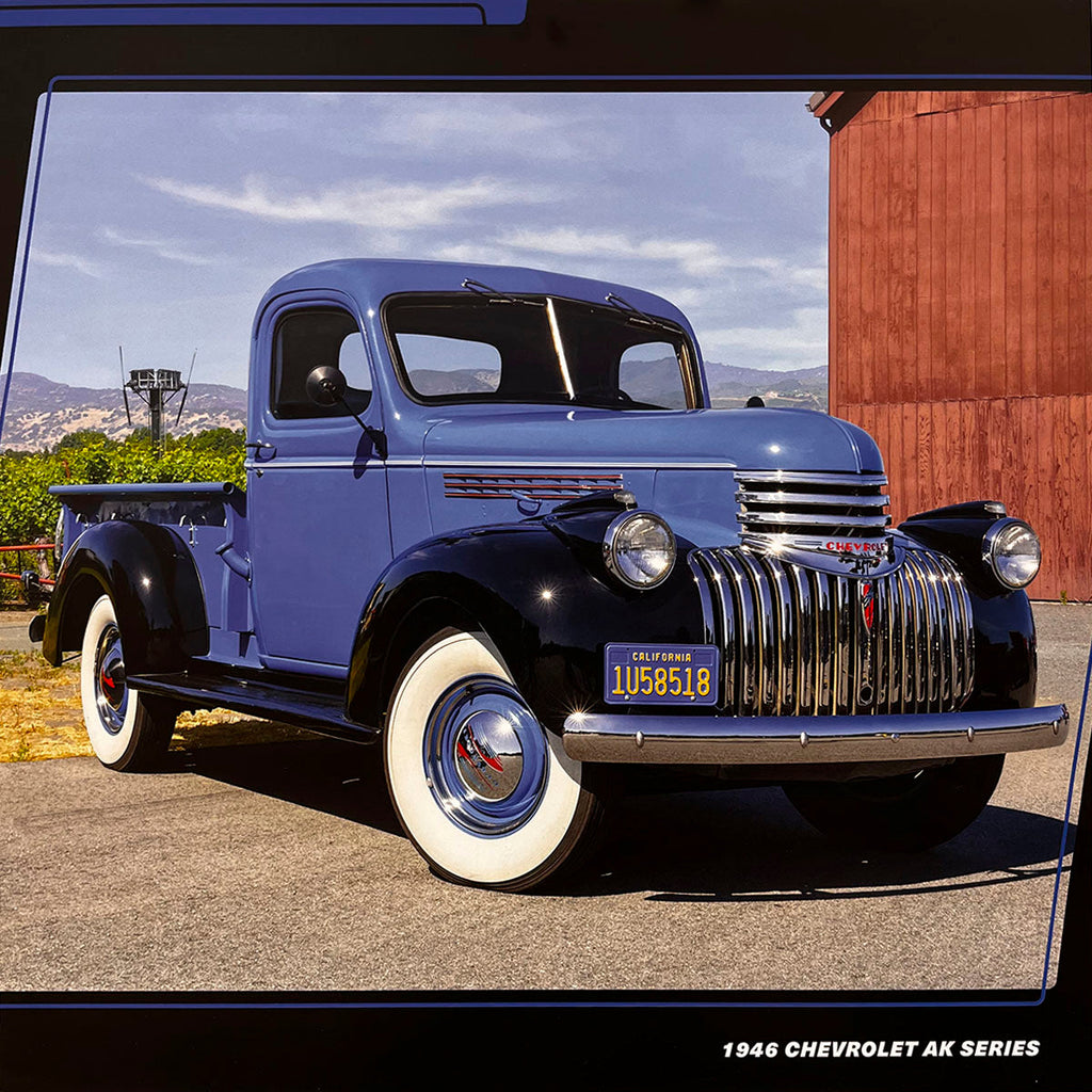 A striking blue and black 1946 Chevrolet AK Series pickup truck with a large chrome grille and white wall tires, parked in an outdoor setting with a red barn in the distance.