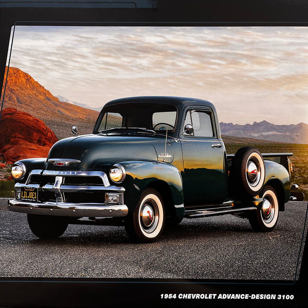 A dark green 1954 Chevrolet Advance-Design 3100 pickup truck with a rear-mounted spare tire and white wall tires, set against a dramatic desert landscape at sunset.