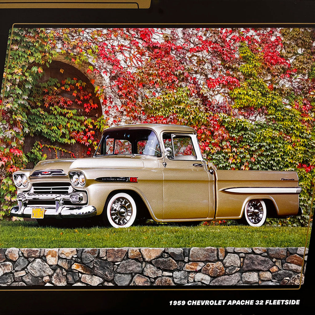 A golden 1959 Chevrolet Apache 32 Fleetside pickup truck with white wall tires, parked on green grass next to a stone wall, with an ivy-covered building behind it.
