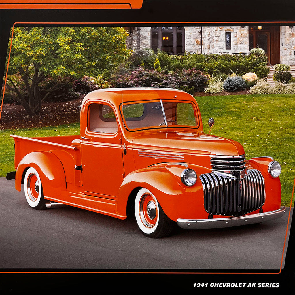 A bright orange 1941 Chevrolet AK Series pickup truck with a large chrome grille and white wall tires, parked on a paved driveway in front of a house with a green lawn.