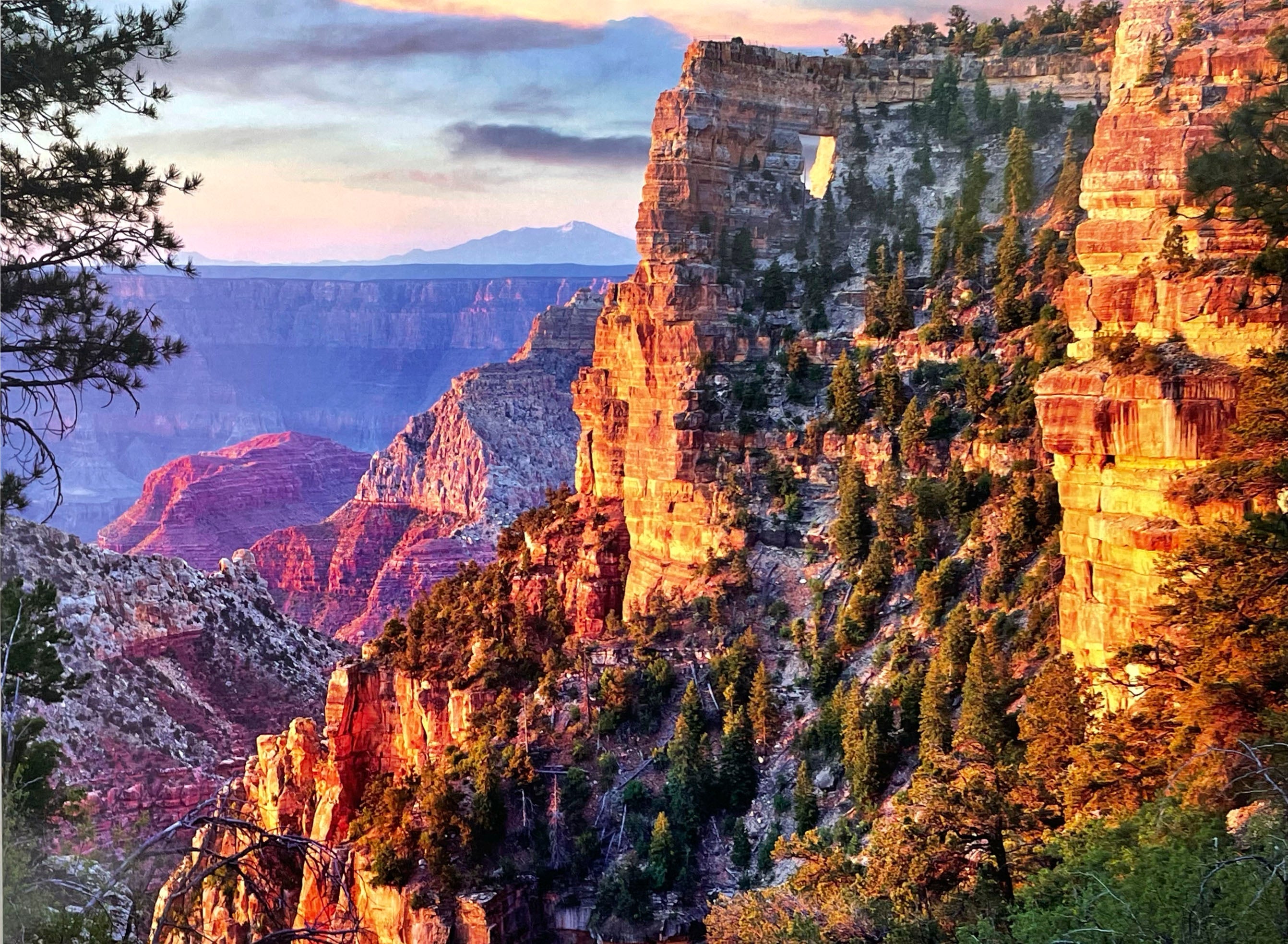 Golden hour lighting on towering Grand Canyon cliffs with scattered pine trees, revealing a natural rock arch against a soft blue sky, an inspiring image for a 2027 calendar.