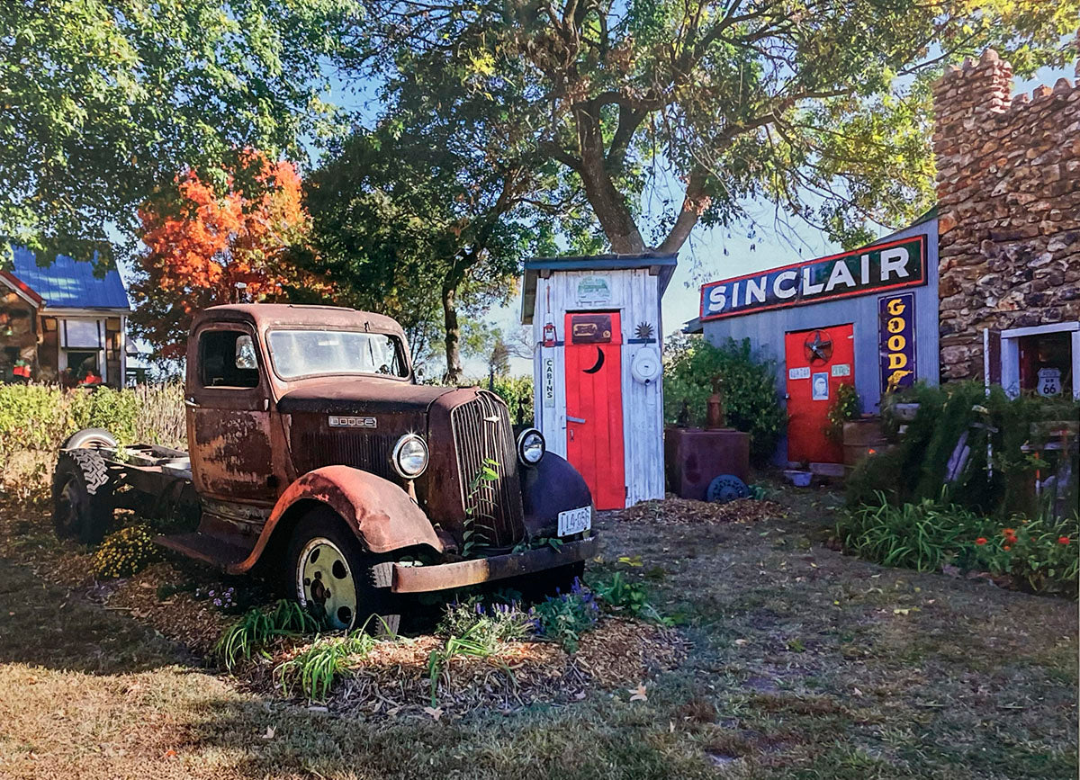 Rustic scene with a weathered vintage Dodge truck parked in front of an old Sinclair Goods store and outhouse.
