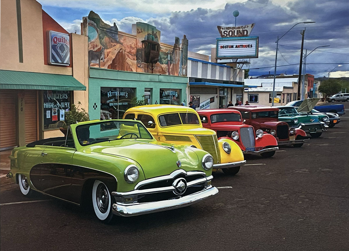 Line of beautifully restored classic cars, including a green convertible, parked along a vibrant Route 66 main street.