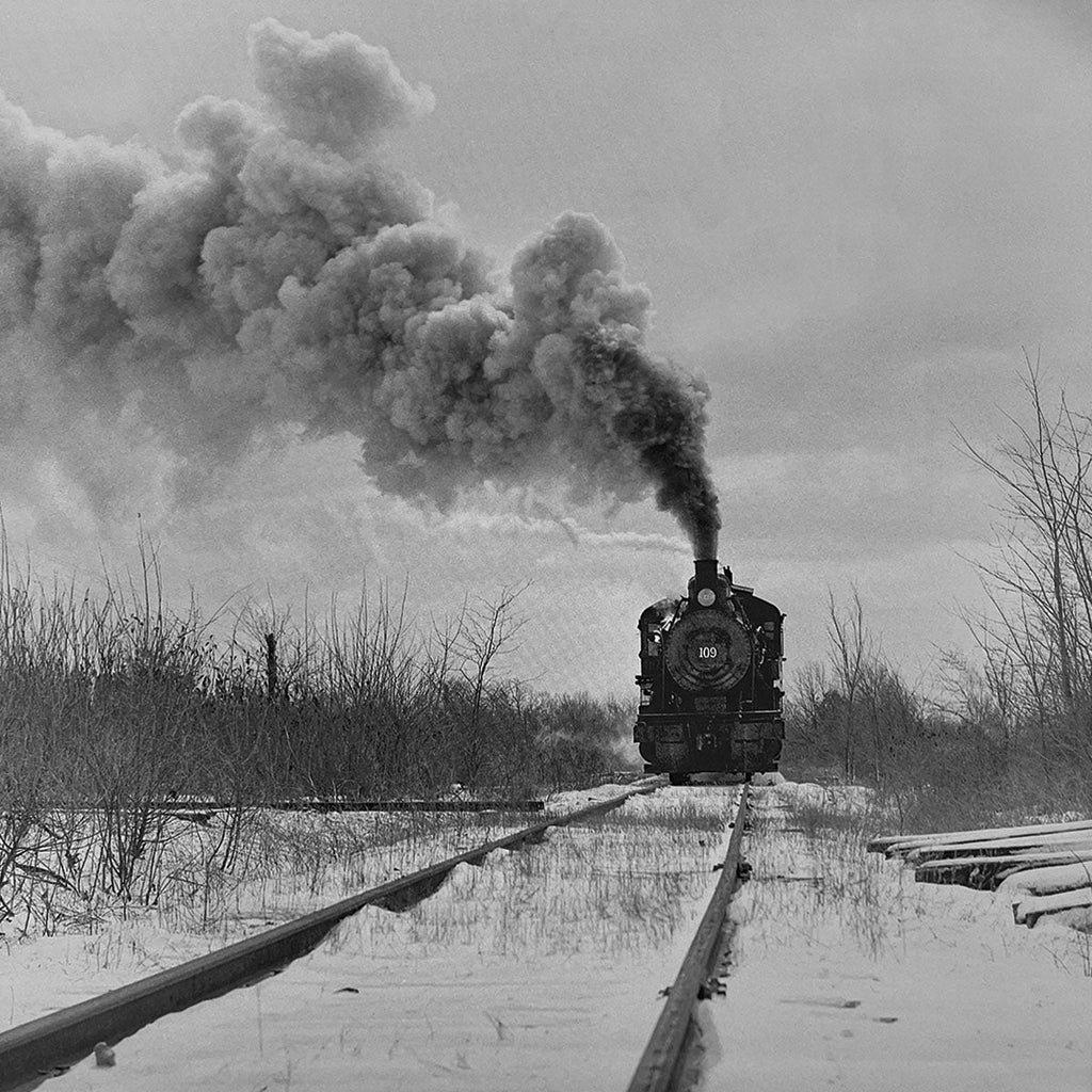 Powerful steam locomotive no. 109 advancing through a snow-covered landscape, emitting thick smoke, vintage black and white railroad photography, winter train scene, classic American railway.