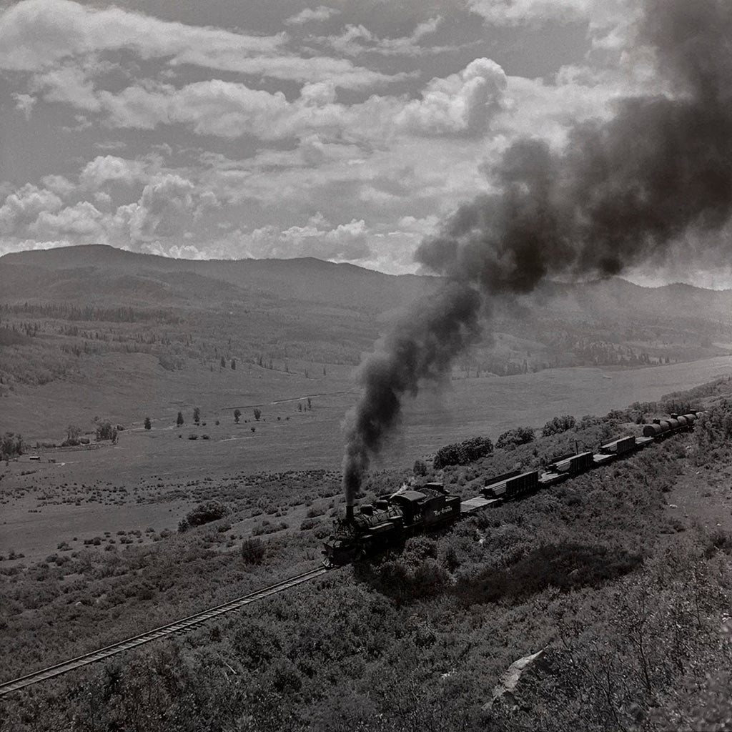 Scenic aerial view of a vintage steam train with freight cars winding along a hillside railway track through a vast, green valley and rolling mountains, historic railroad photography.