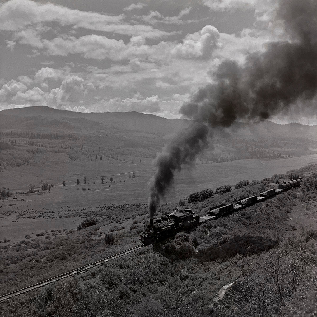 Scenic aerial view of a vintage steam train with freight cars winding along a hillside railway track through a vast, green valley and rolling mountains, historic railroad photography.