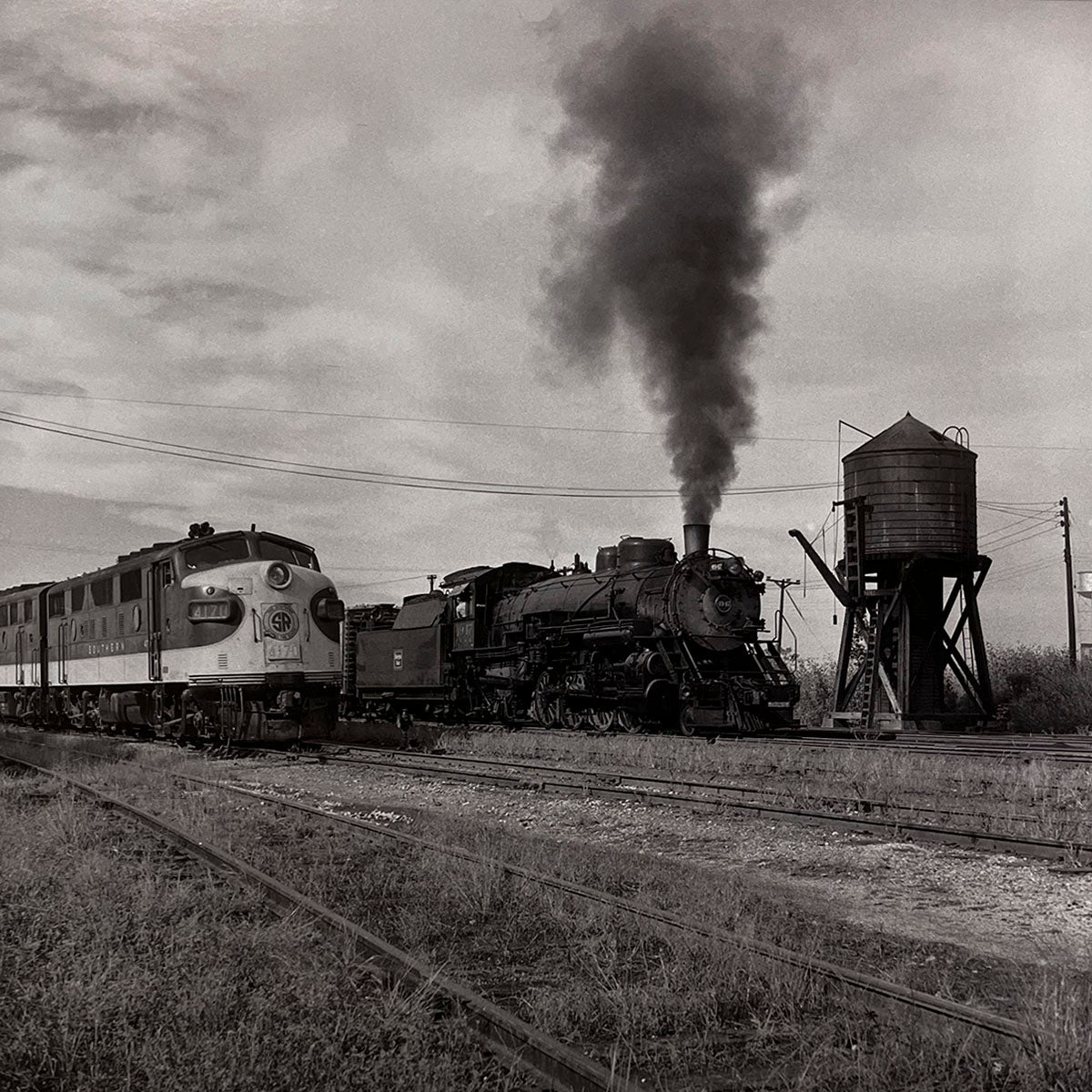 Black and white photo showing a classic steam locomotive beside a modern diesel train, with a rustic water tower and multiple railway tracks in an industrial setting, railroad history.