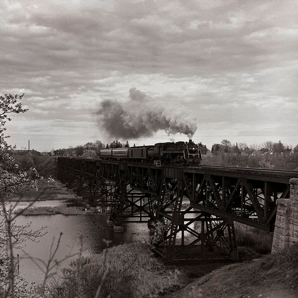 Majestic steam passenger train crossing a large, intricate steel truss bridge spanning a wide river with trees on the banks, captured in timeless black and white, impressive railway infrastructure.
