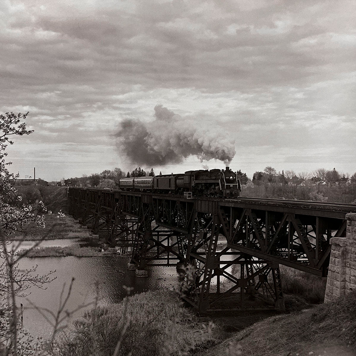 Majestic steam passenger train crossing a large, intricate steel truss bridge spanning a wide river with trees on the banks, captured in timeless black and white, impressive railway infrastructure.