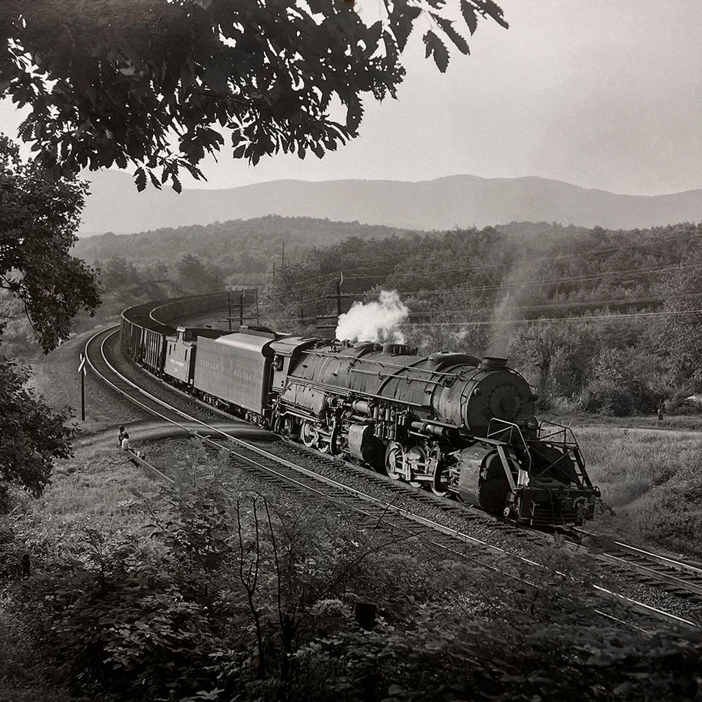 Long steam freight train curves through a scenic rural landscape, dense foliage framing the tracks and distant hills under a cloudy sky, classic American steam era.