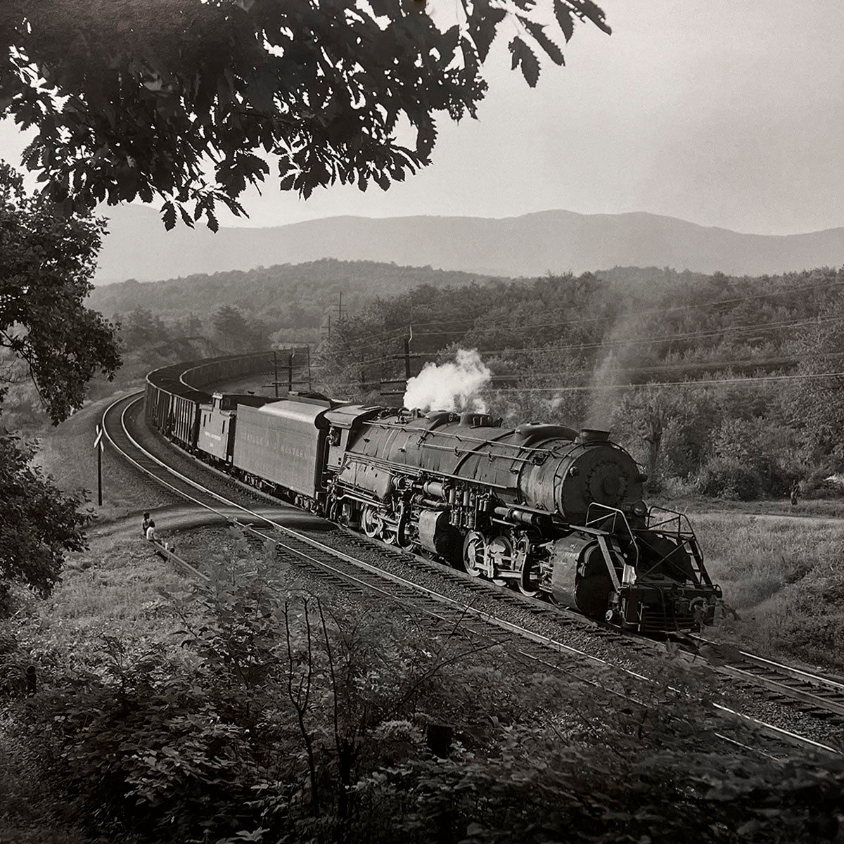 Long steam freight train curves through a scenic rural landscape, dense foliage framing the tracks and distant hills under a cloudy sky, classic American steam era.