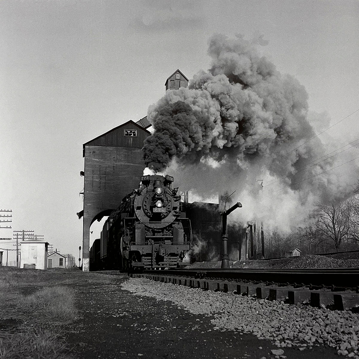 Dramatic black and white image of a steam locomotive emerging from an industrial building or shed, clouds of smoke pouring from its stack, active rail yard scene, heavy industry.