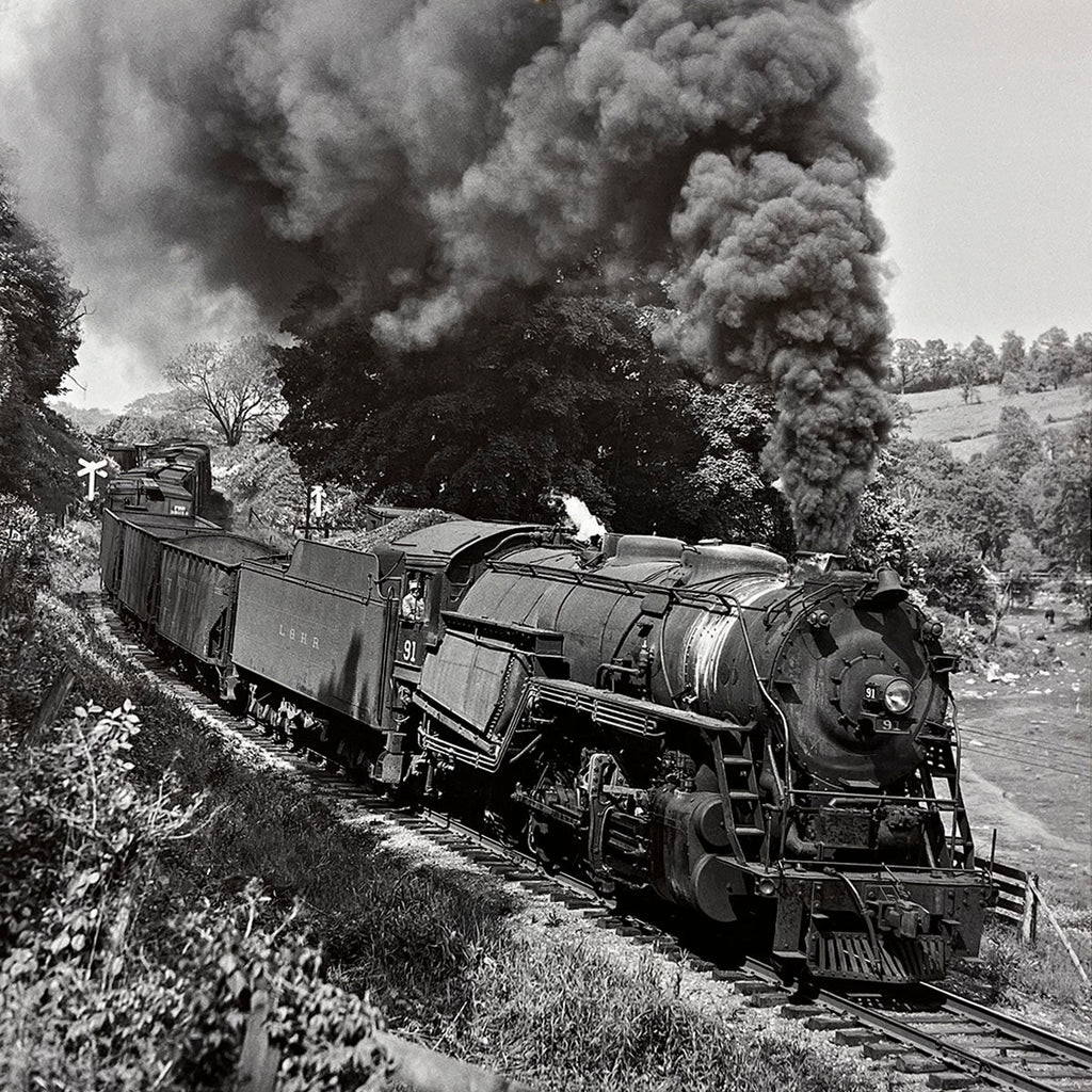 Close-up of a large vintage steam engine number 91 with freight cars climbing a gentle slope, surrounded by lush vegetation and rolling hills, classic railroading.