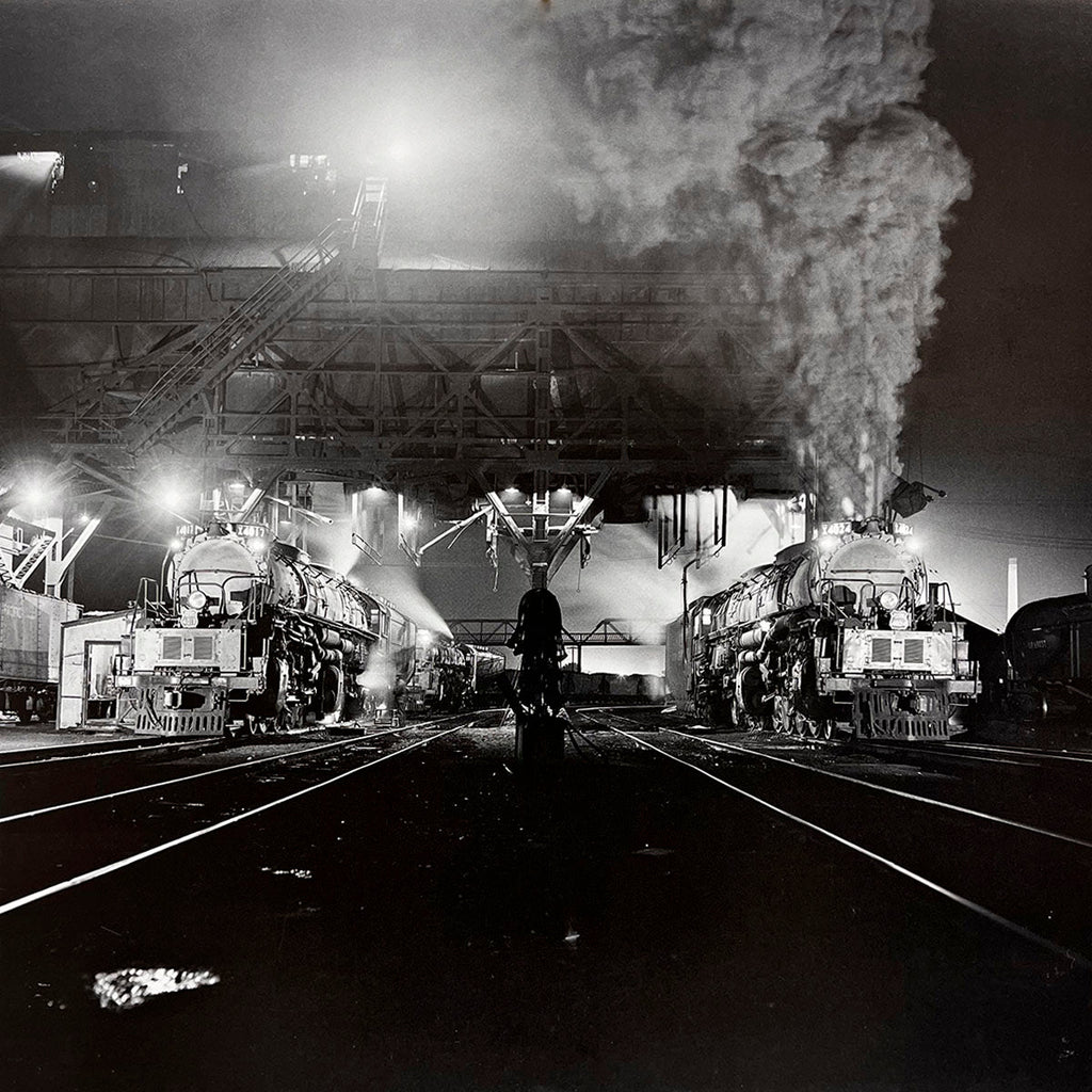 Striking night scene of two illuminated steam locomotives steaming in a busy train yard, atmospheric lighting highlighting industrial structures, powerful engines at rest.