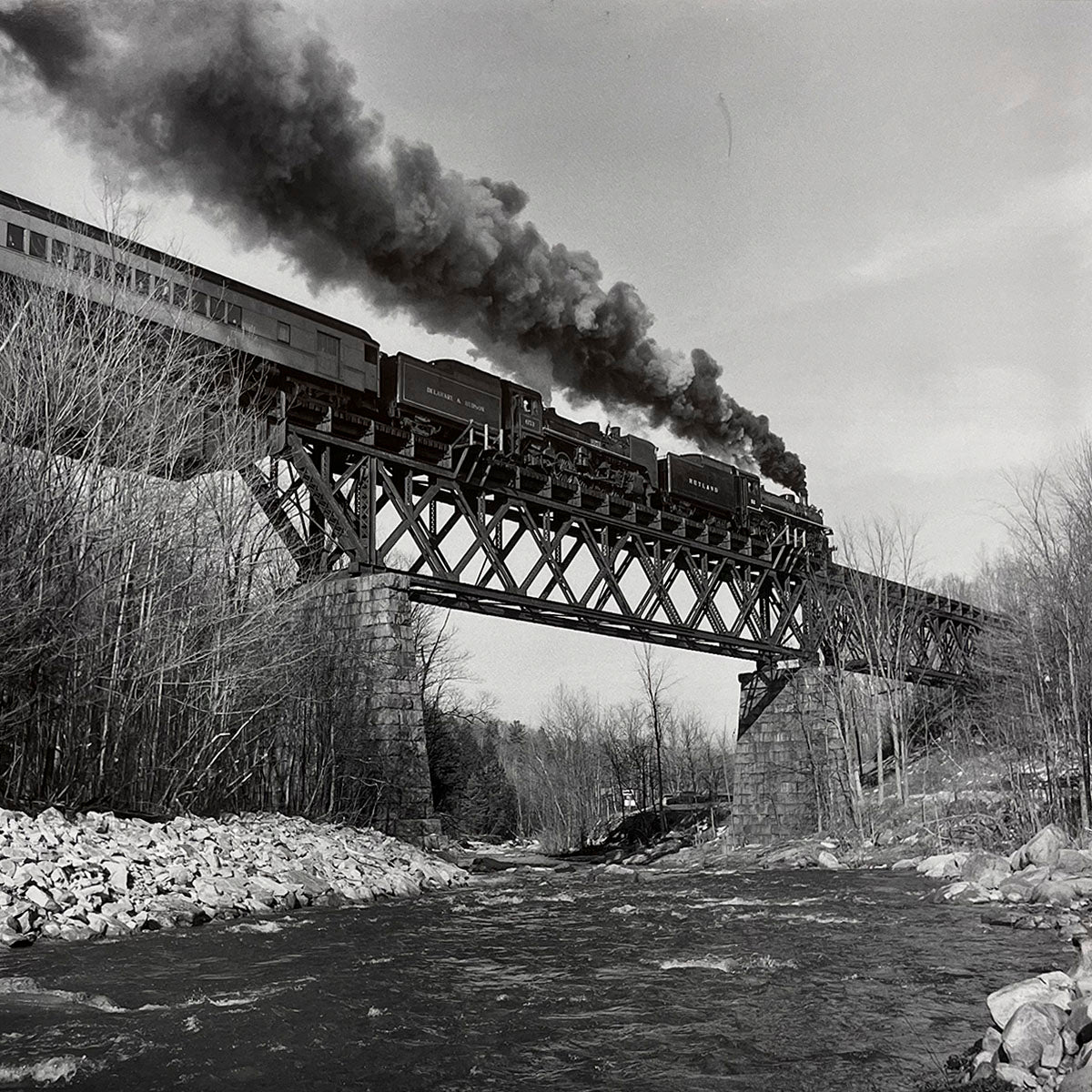 Passenger steam train crossing a high, sturdy metal trestle bridge over a river, surrounded by bare winter trees, an iconic view of railway engineering and travel.