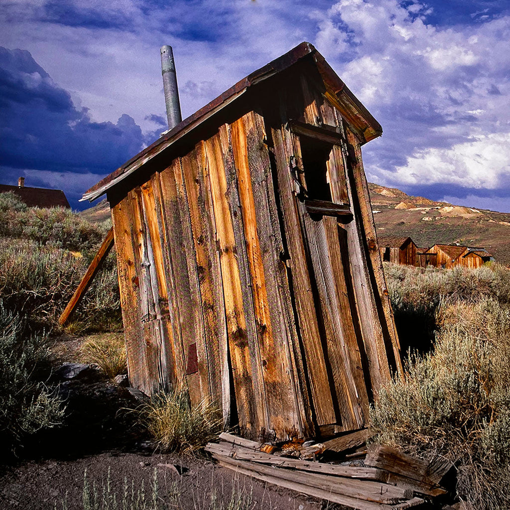 March 2027 calendar image: A leaning, weathered wooden outhouse with a tin roof and open window, amidst dry brush in a desert-like landscape, with distant hills under a dramatic sky.