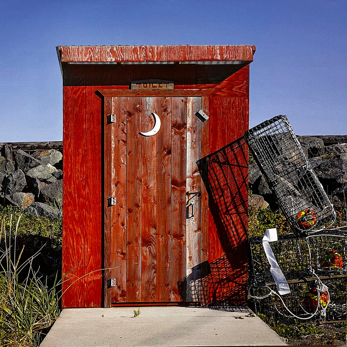 April 2027 calendar image: A vibrant red outhouse with a classic crescent moon cutout on the door, standing on concrete next to lobster traps, against a rock wall and clear blue sky.