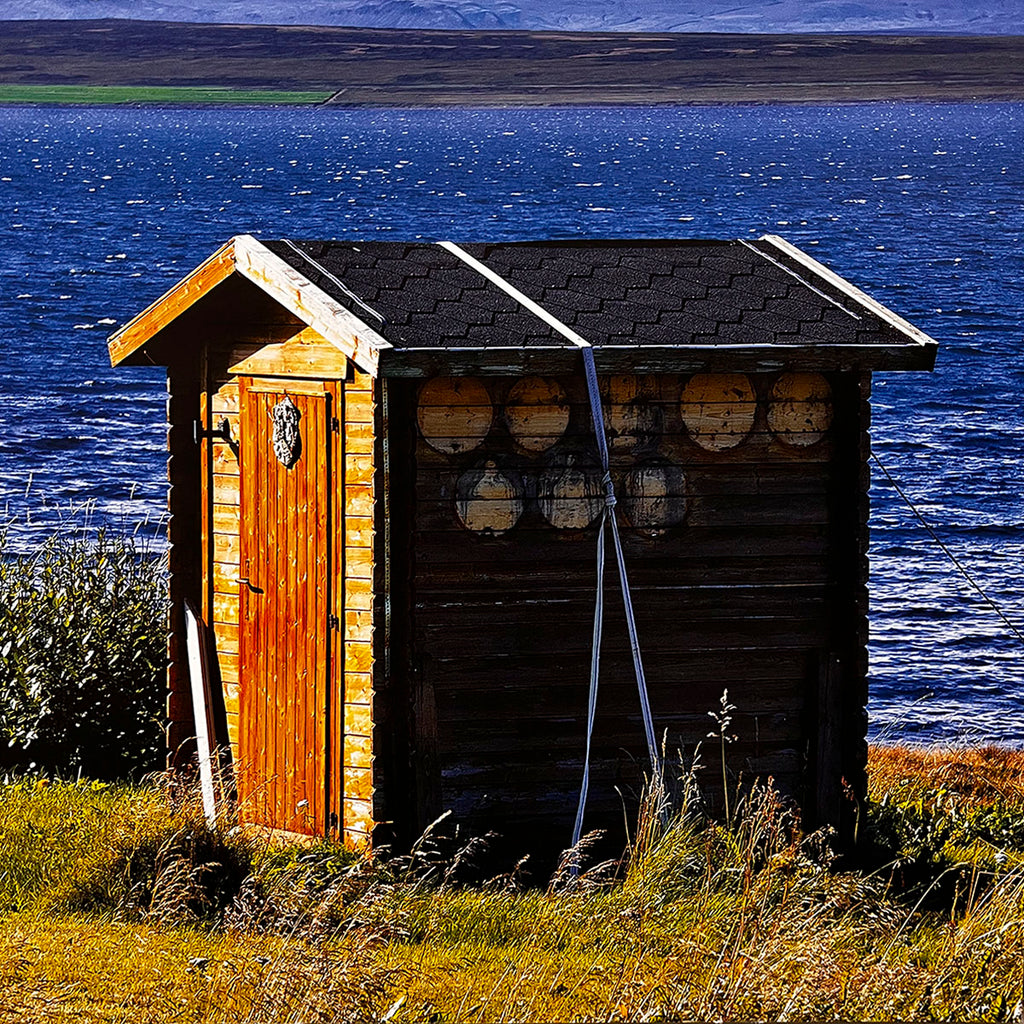 May 2027 calendar image: A sturdy wooden outhouse with a black shingle roof and decorative accents, positioned by the clear blue waters of a lake with a distant shoreline.