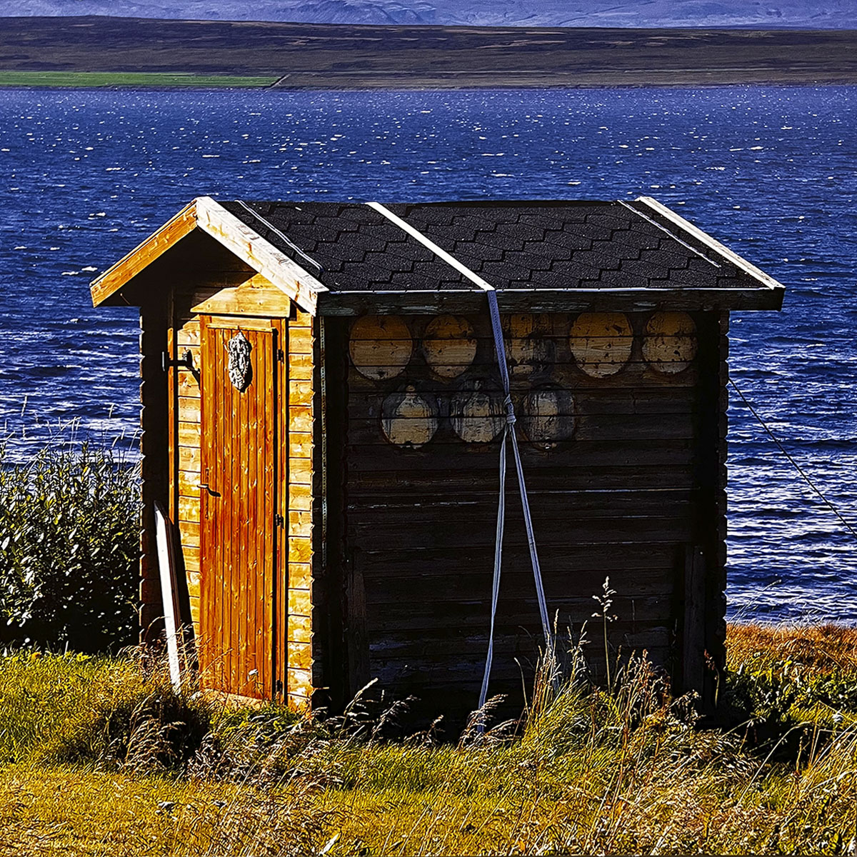May 2027 calendar image: A sturdy wooden outhouse with a black shingle roof and decorative accents, positioned by the clear blue waters of a lake with a distant shoreline.