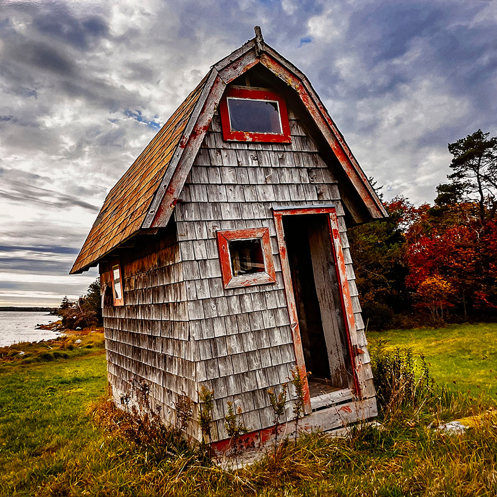 July 2027 calendar image: A rustic, leaning wooden outhouse with red window frames and trim, situated in a green grassy field overlooking a body of water and autumn-colored trees.