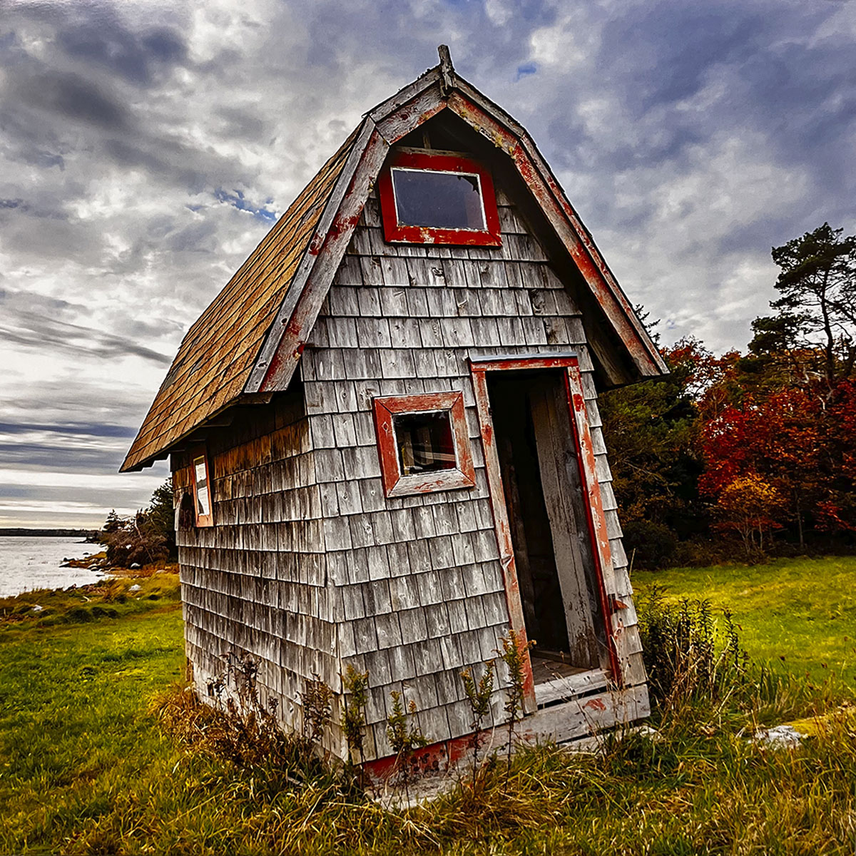 July 2027 calendar image: A rustic, leaning wooden outhouse with red window frames and trim, situated in a green grassy field overlooking a body of water and autumn-colored trees.