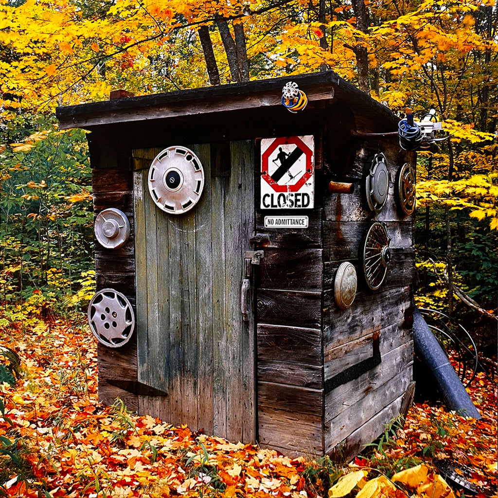 September 2027 calendar image: A creatively decorated wooden outhouse with a 'CLOSED NO ADMITTANCE' sign, hubcaps, and other items affixed to its exterior, surrounded by colorful autumn leaves.