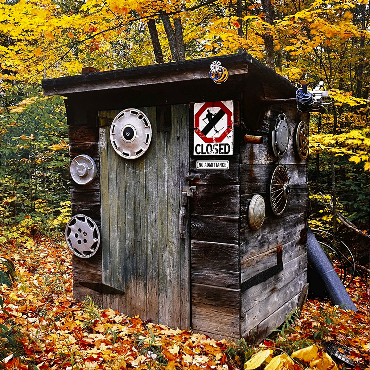 September 2027 calendar image: A creatively decorated wooden outhouse with a 'CLOSED NO ADMITTANCE' sign, hubcaps, and other items affixed to its exterior, surrounded by colorful autumn leaves.