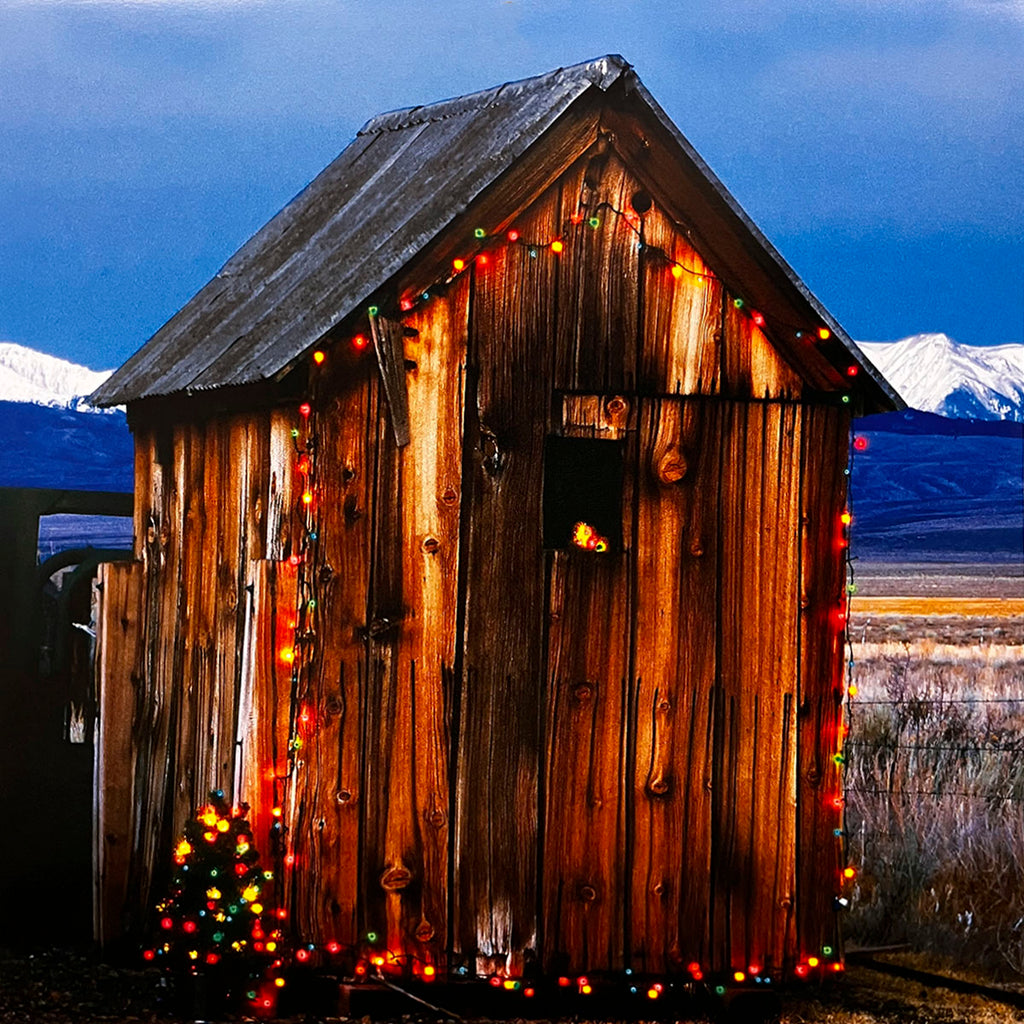 October 2027 calendar image: A weathered wooden outhouse adorned with festive colorful Christmas lights, set against a dark blue sky with snow-capped mountains in the distant background.