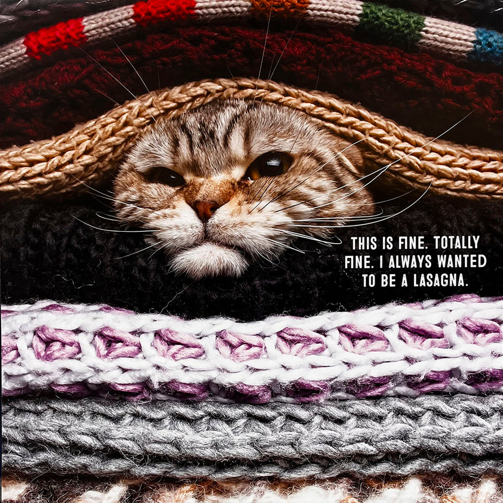 Hilarious cat calendar image showing a tabby cat's face peering out from under a stack of colorful knitted blankets. Caption says: 'This is fine. Totally fine. I always wanted to be a lasagna.'