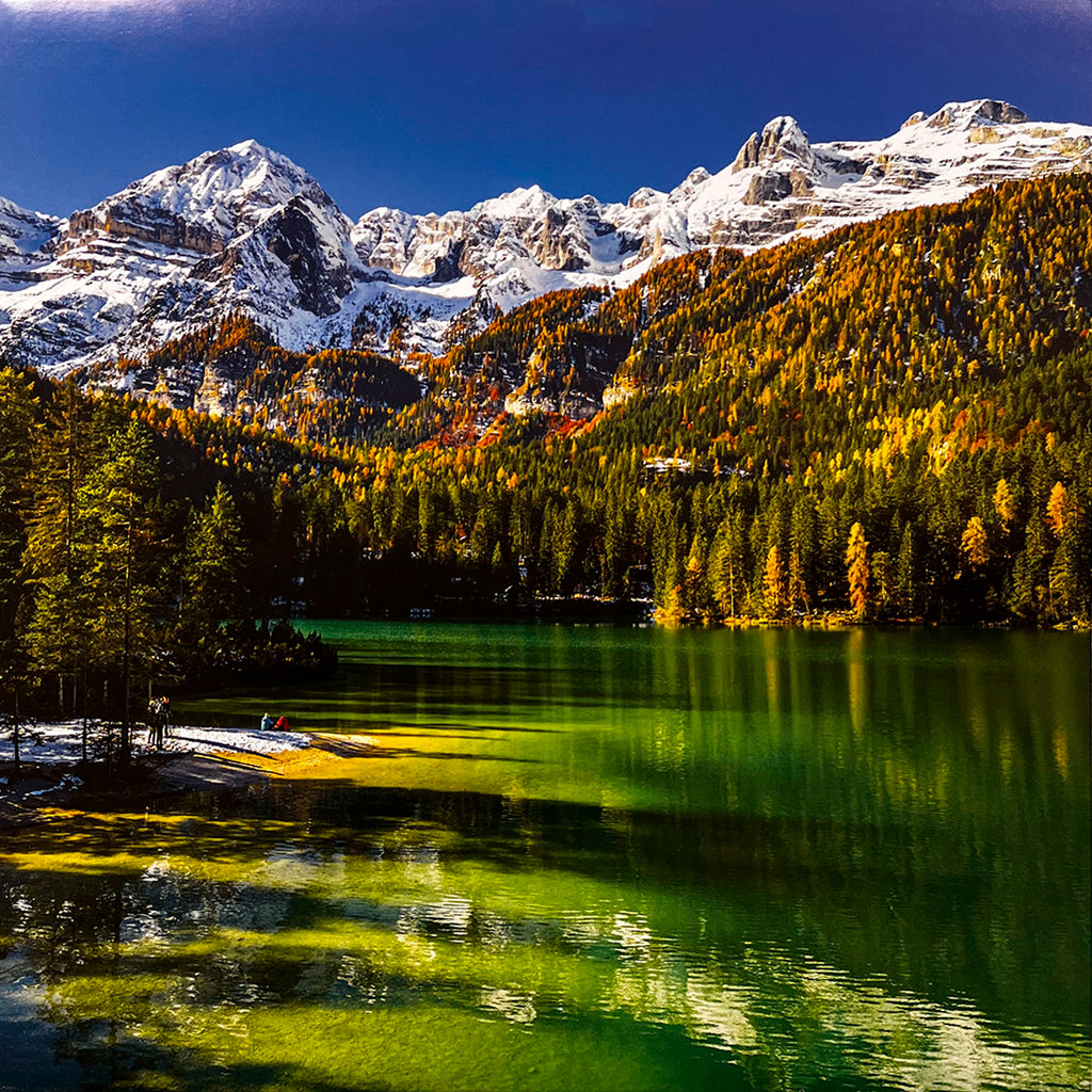 Breathtaking emerald-green lake nestled within an autumn forest with vibrant yellow and orange trees, backed by majestic snow-capped mountains.