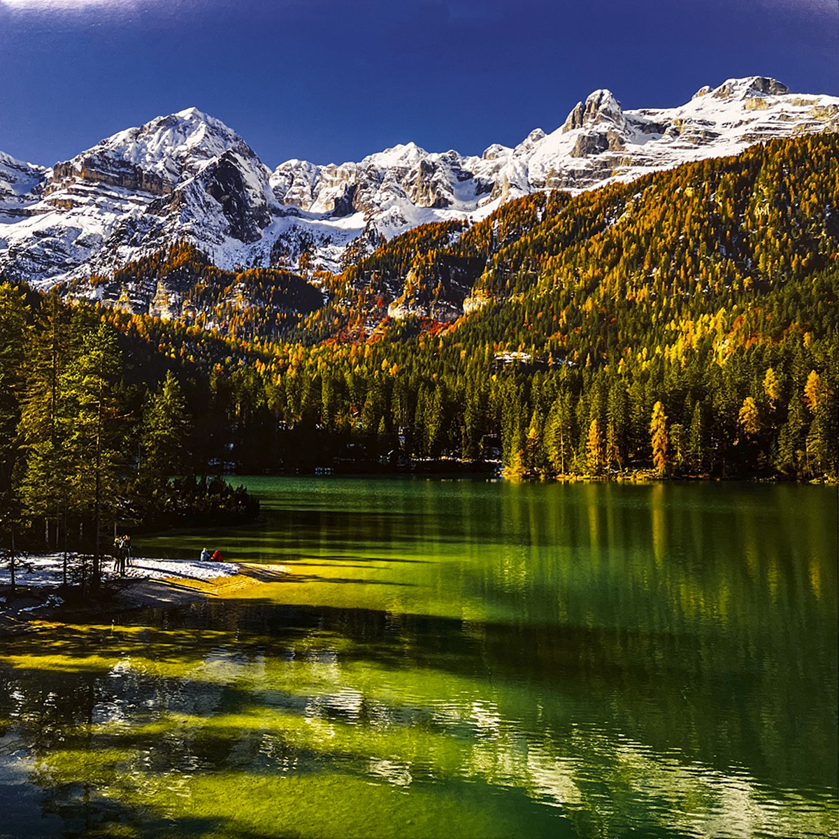 Breathtaking emerald-green lake nestled within an autumn forest with vibrant yellow and orange trees, backed by majestic snow-capped mountains.