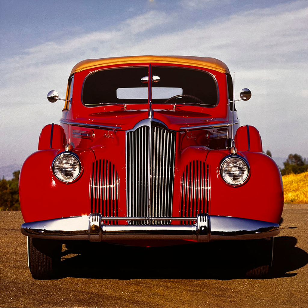 The bold front view of a gleaming red 1941 Packard Super Eight convertible, showcasing its iconic waterfall grille and elegant chrome bumper under a clear sky, a true luxury classic car.