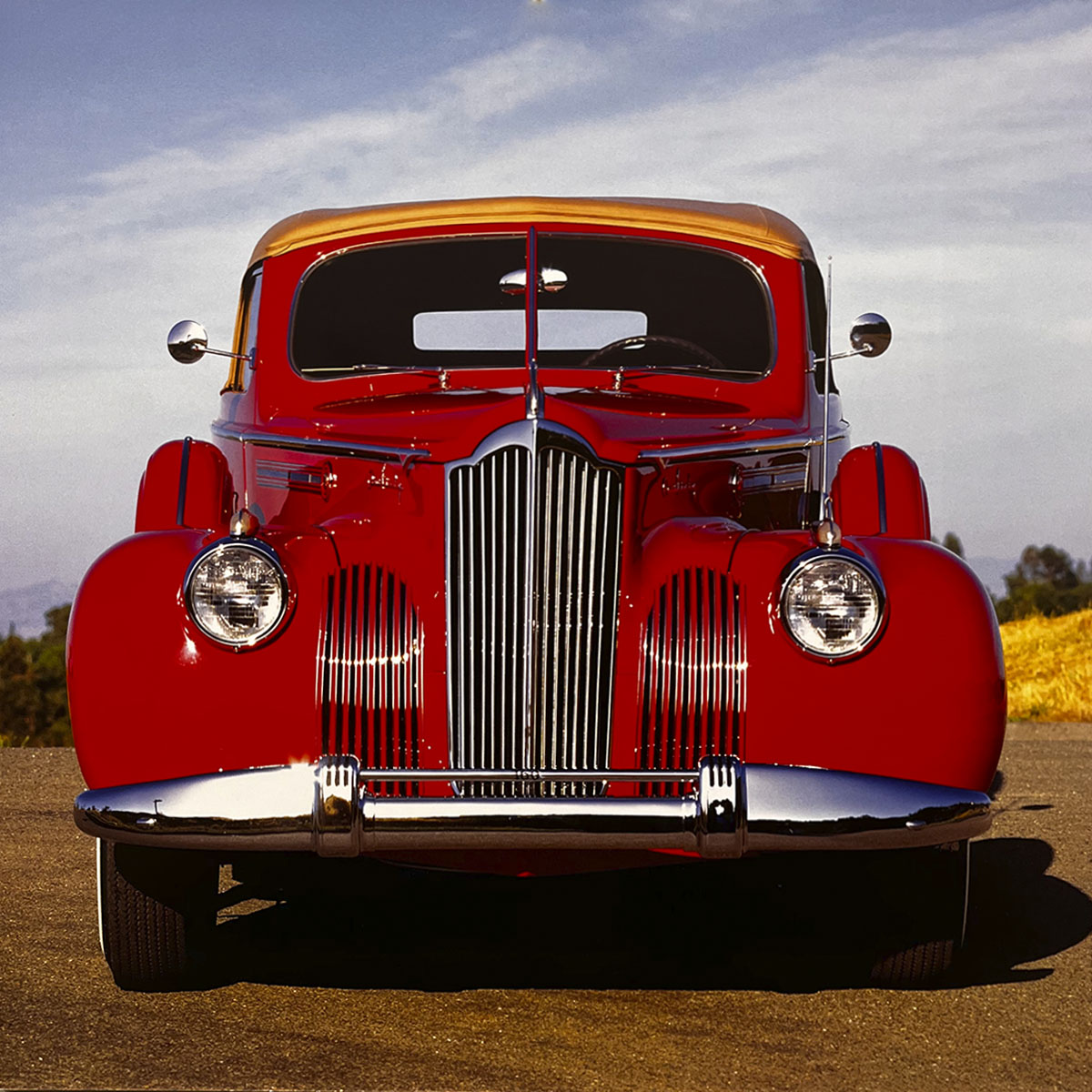 The bold front view of a gleaming red 1941 Packard Super Eight convertible, showcasing its iconic waterfall grille and elegant chrome bumper under a clear sky, a true luxury classic car.