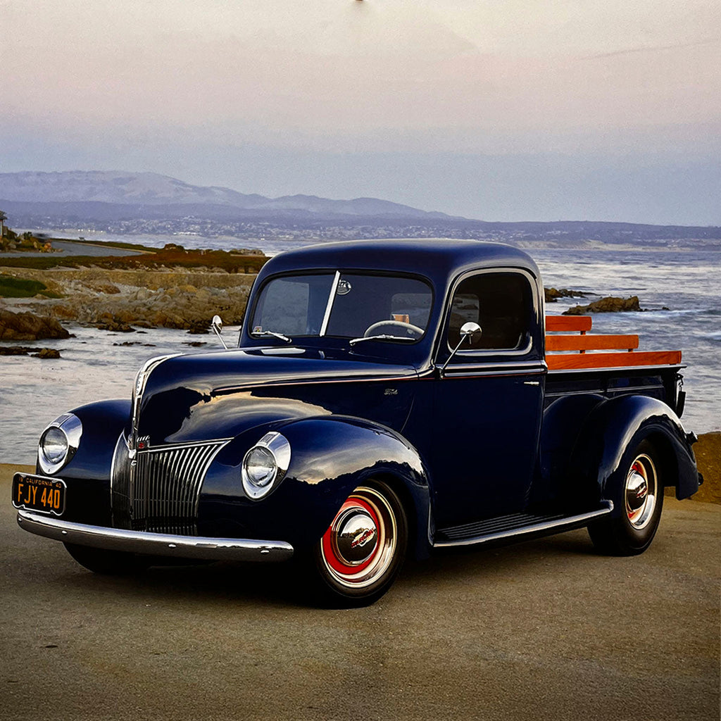 A deep navy blue 1940 Ford pickup truck with wooden bed rails, parked by a scenic coastline with the ocean and distant hills, epitomizing classic American utility vehicles.