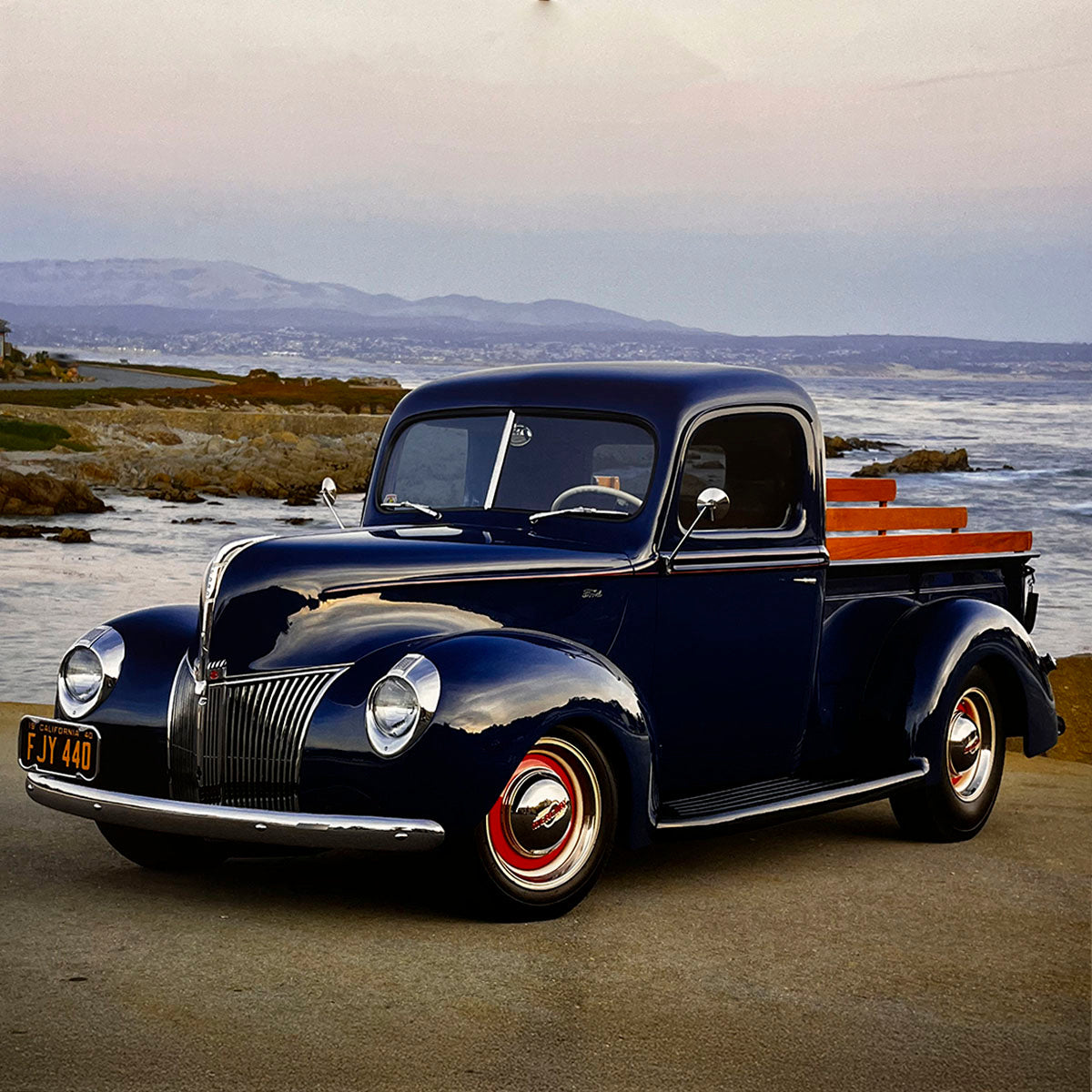 A deep navy blue 1940 Ford pickup truck with wooden bed rails, parked by a scenic coastline with the ocean and distant hills, epitomizing classic American utility vehicles.