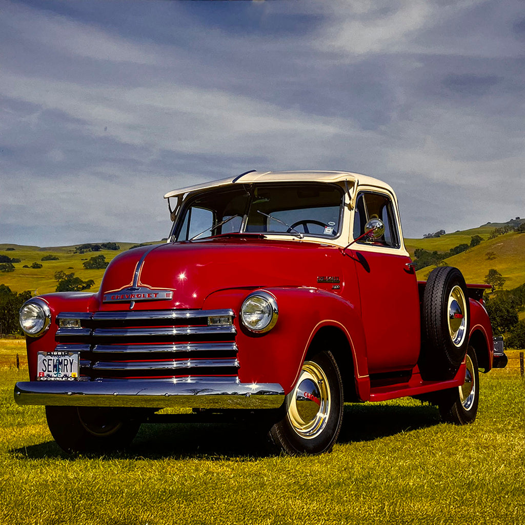 A striking red and cream two-tone 1951 Chevrolet 3100 pickup truck, parked in a lush green field under a partly cloudy sky, featuring a side-mounted spare tire and classic styling.